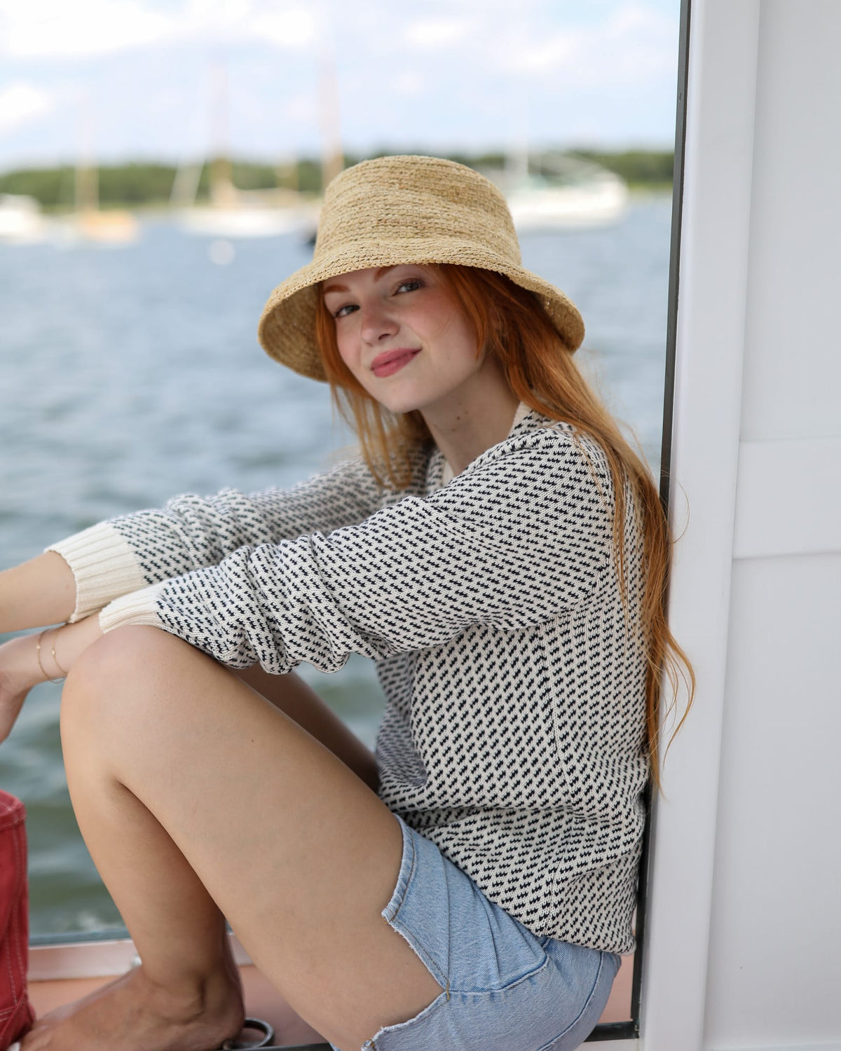 Whalerknits Sakonnet Birdseye Crewneck Pullover: A woman in natural with navy birdseye pattern, denim shorts and a straw hat sits aboard a boat.