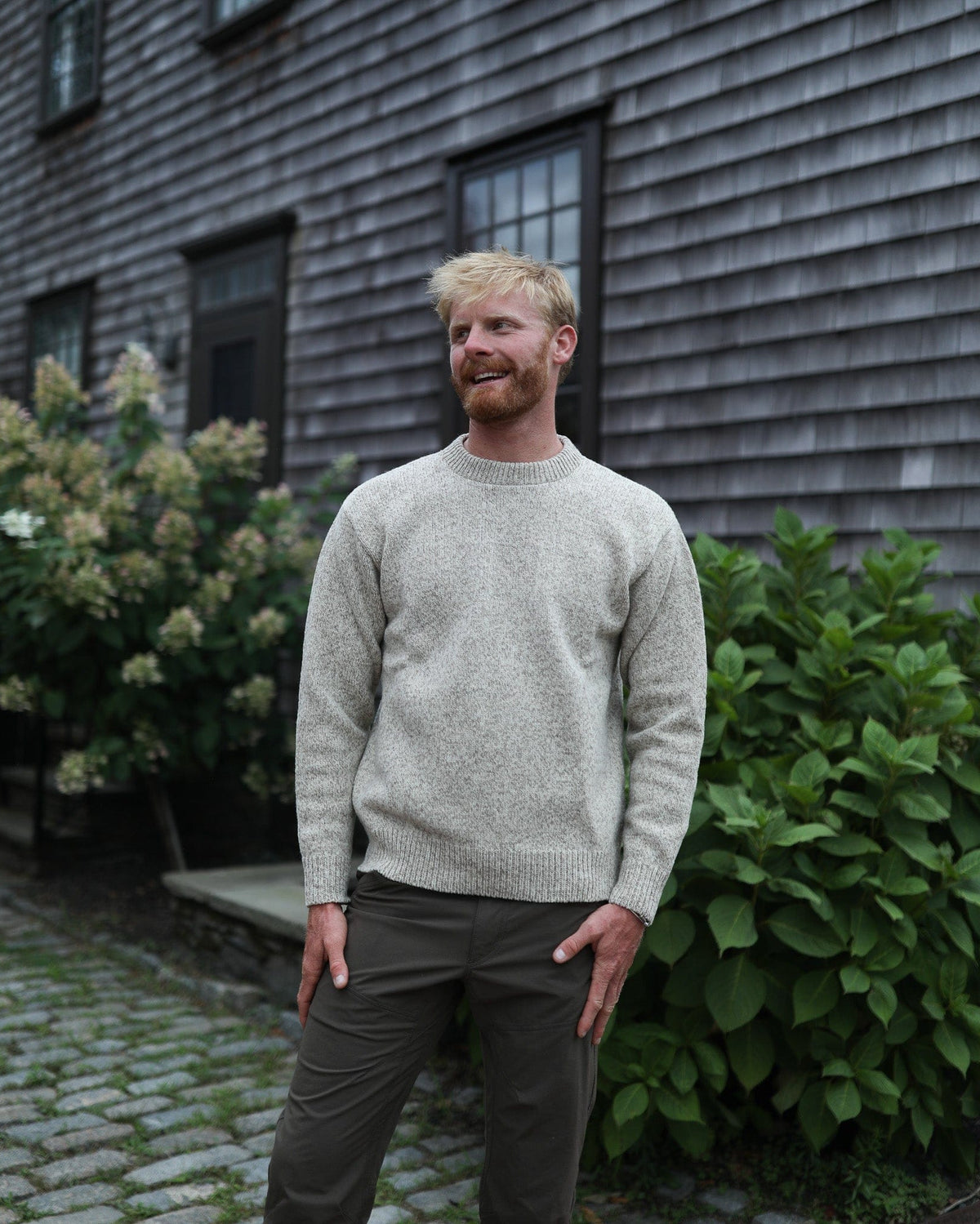 Whalerknits Wellfleet Classic Crewneck Jersey - Wool Blend: A man in the light heathered sweater stands beside a shingled wall and shrubs.