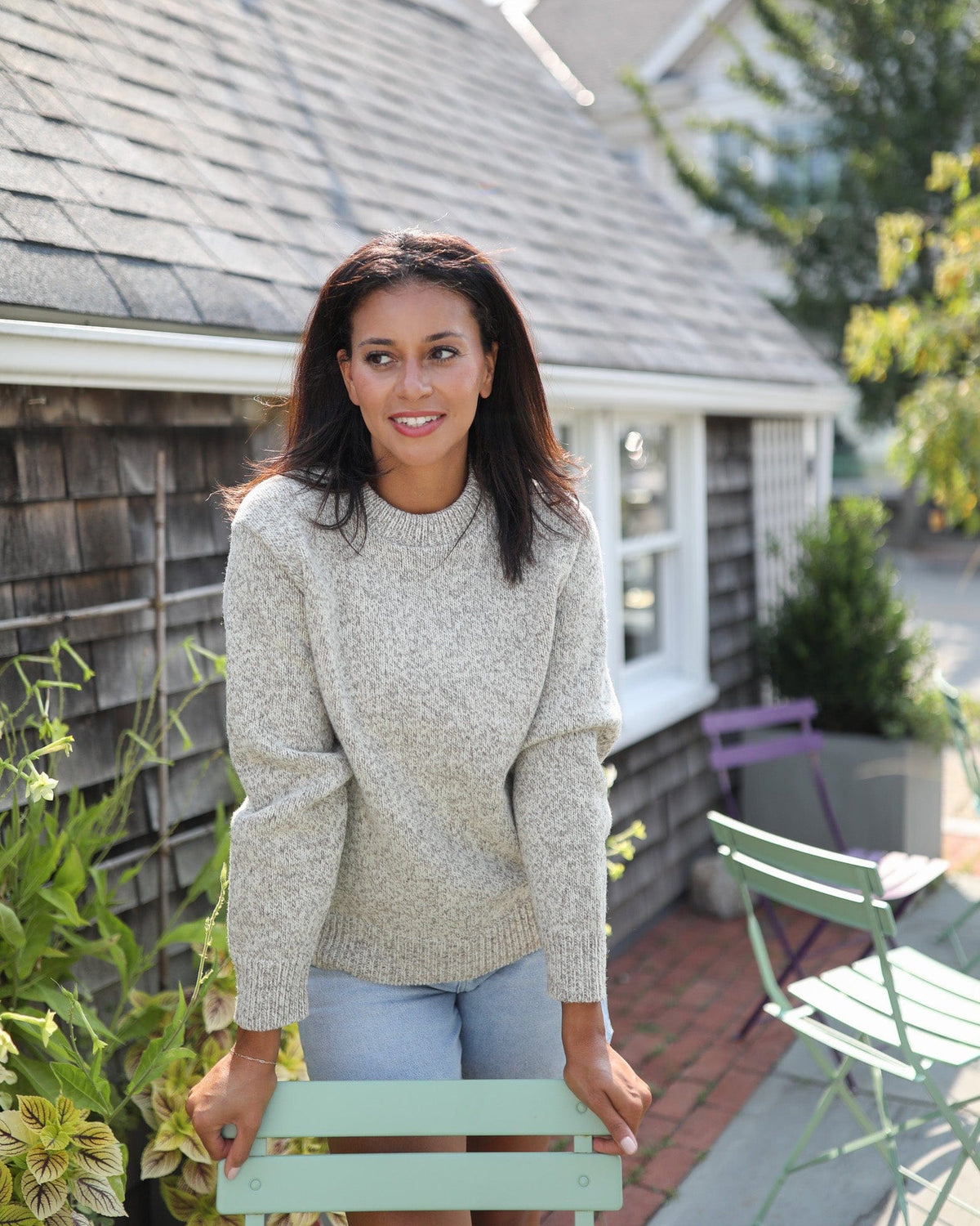 Whalerknits Wellfleet Classic Crewneck Jersey - Wool Blend: A woman wears the light heathered sweater with jeans beside a green chair outside a shingled building.