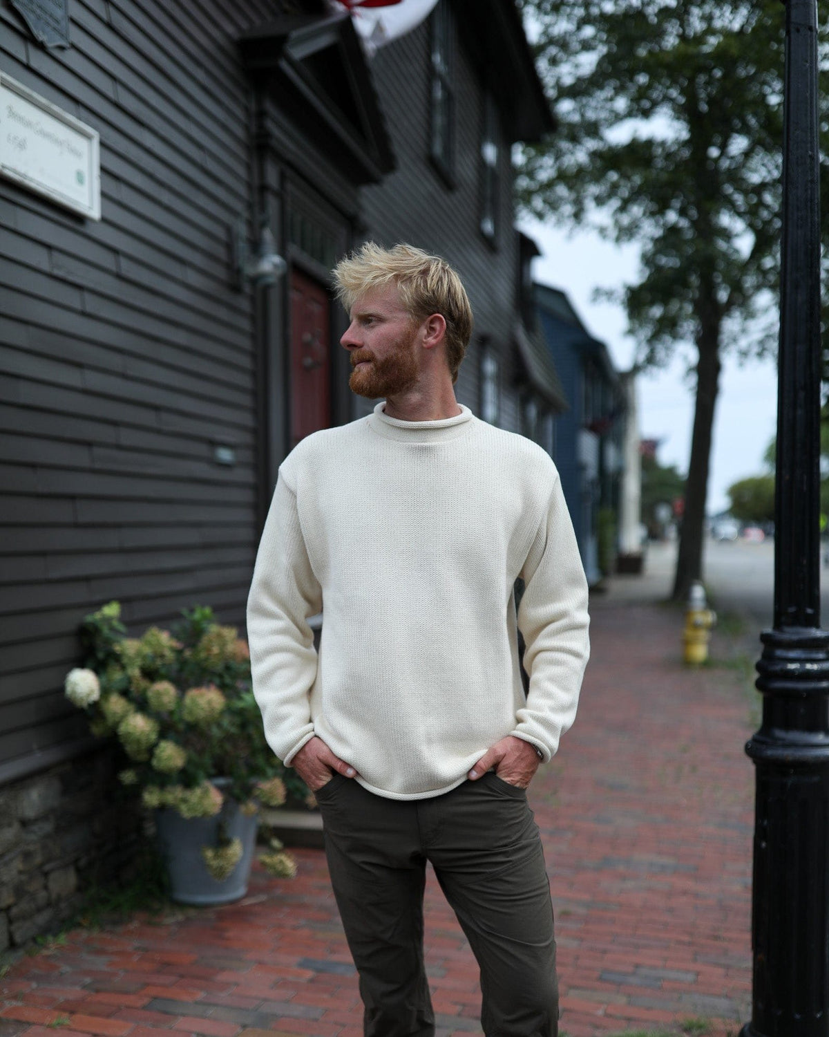 Whalerknits Wellfleet Rollneck Sweater: A man wears natural with dark trousers on a brick sidewalk beside a shingled building.