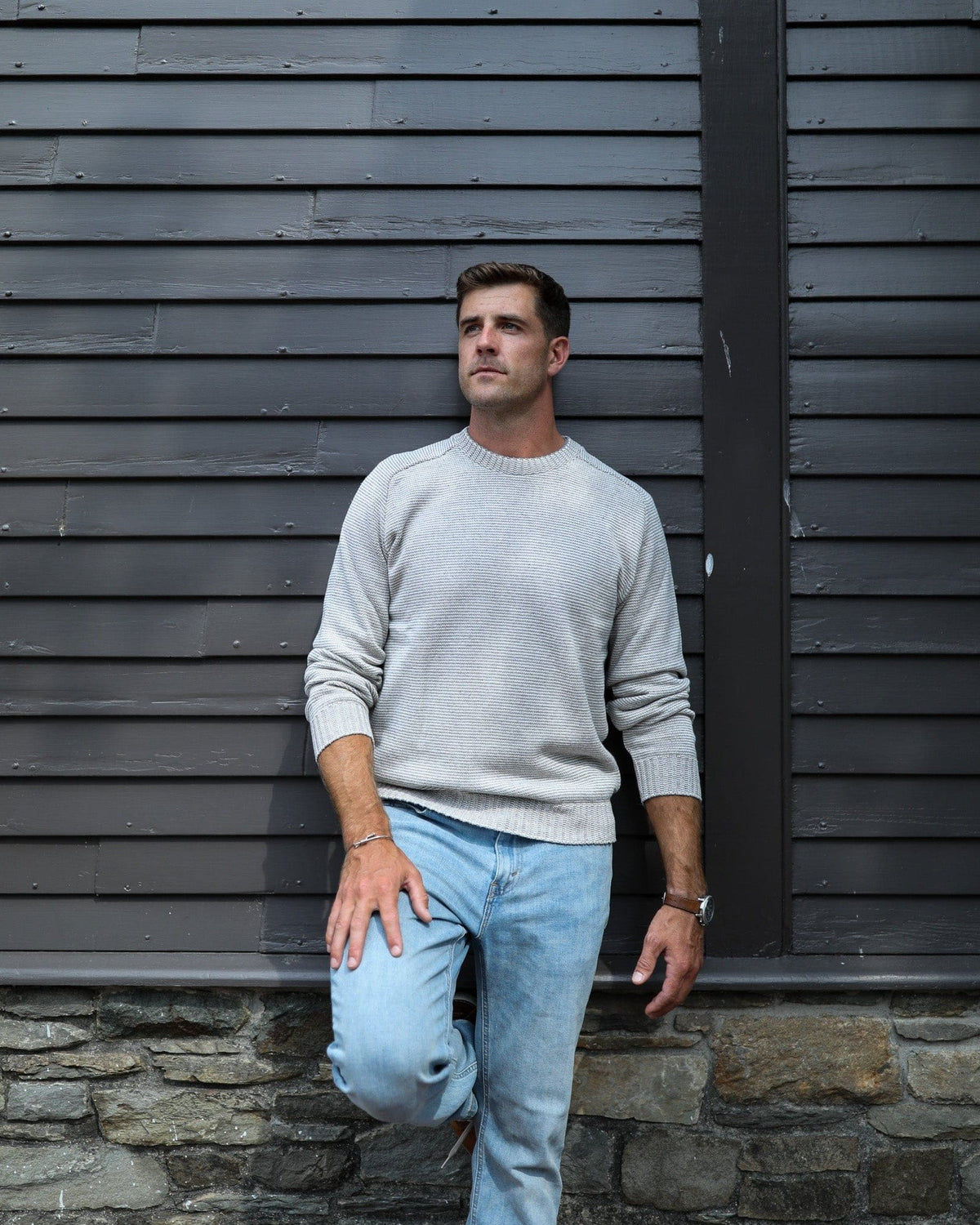 Whalerknits Westport Crewneck Saddle Shoulder Pullover: A man wears the light fine-striped sweater with jeans against a dark clapboard wall.