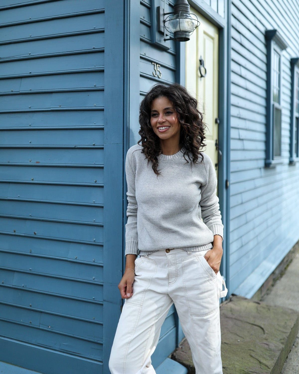Whalerknits Westport Crewneck Saddle Shoulder Pullover: A woman in the light fine-striped sweater and white trousers stands beside a blue house.