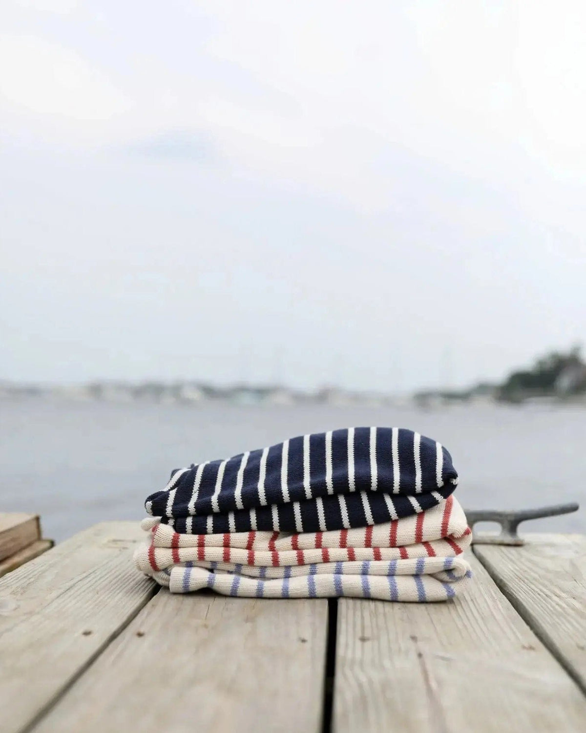 Whalerknits Nantucket Classic Boatneck Sweater: Three folded striped sweaters stacked on a wooden dock beside the water.