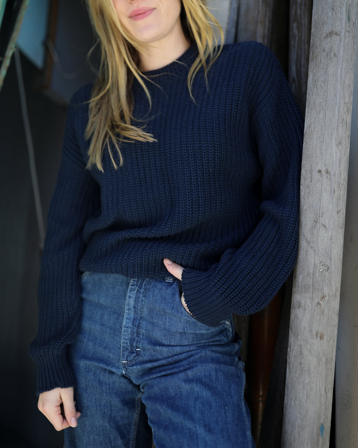 Whalerknits Newport Shaker Knit Crewneck Sweater: Close view of a woman wearing navy with jeans, one hand in a pocket beside a wooden post.